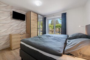 Une chambre à coucher avec un lit, un téléviseur fixé au mur, une commode en bois et de grandes fenêtres.