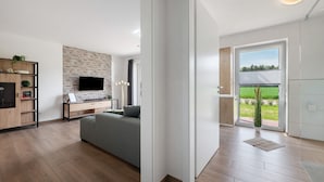 A modern living room with a flat-screen TV, a grey sofa, wooden flooring, a stone wall, and a view of a green landscape through a glass door. - Holiday Apartment 'Anna 1 Eg' with Private Terrace and Garden (Suhle)