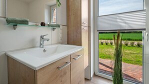 A modern bathroom with a large mirror, a white sink, wooden cabinets, and a view of a green landscape through a glass door. - Holiday Apartment 'Anna 1 Eg' with Private Terrace and Garden (Suhle)