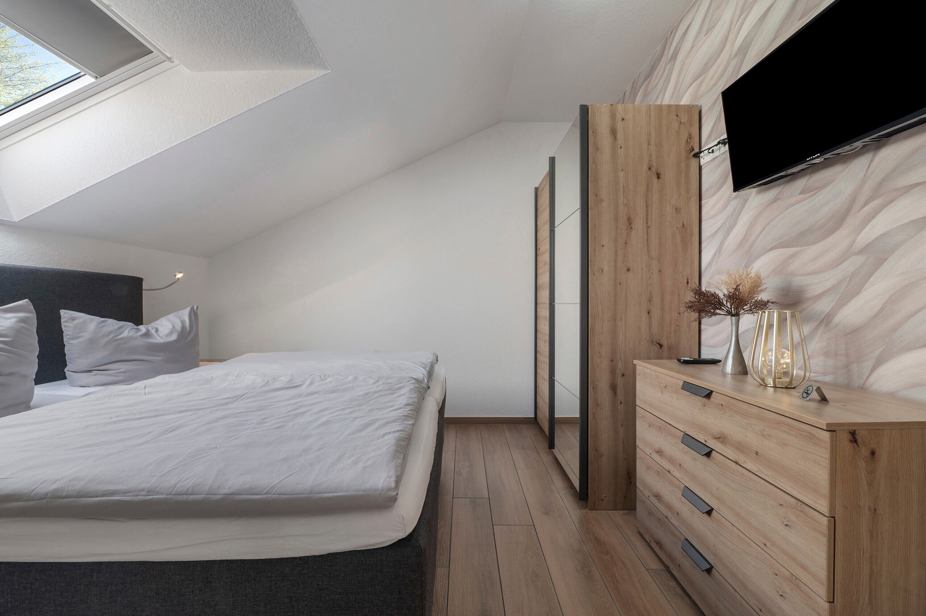 Ein Schlafzimmer mit einem Bett, einer Holzkommode, einem an der Wand befestigten Fernseher und einem Dachfenster.