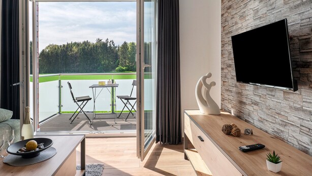 Ein modernes Wohnzimmer mit großem Fernseher, einem Holztisch, einer kleinen Pflanze und einem Balkon mit Tisch und Stühlen.