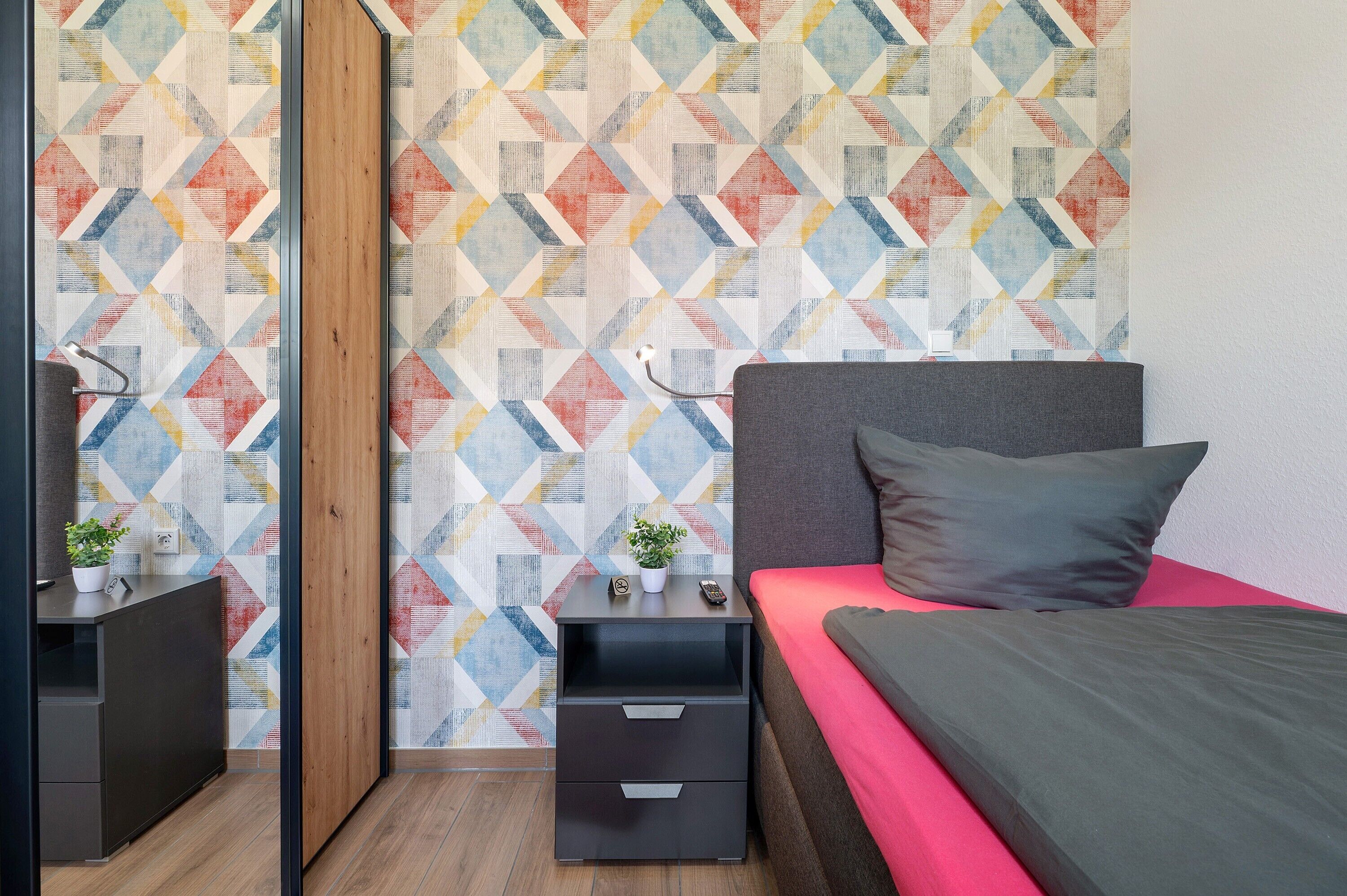 A modern bedroom with a geometric-patterned wallpaper, a bed with a gray headboard, a nightstand with drawers, and a small plant on the nightstand.