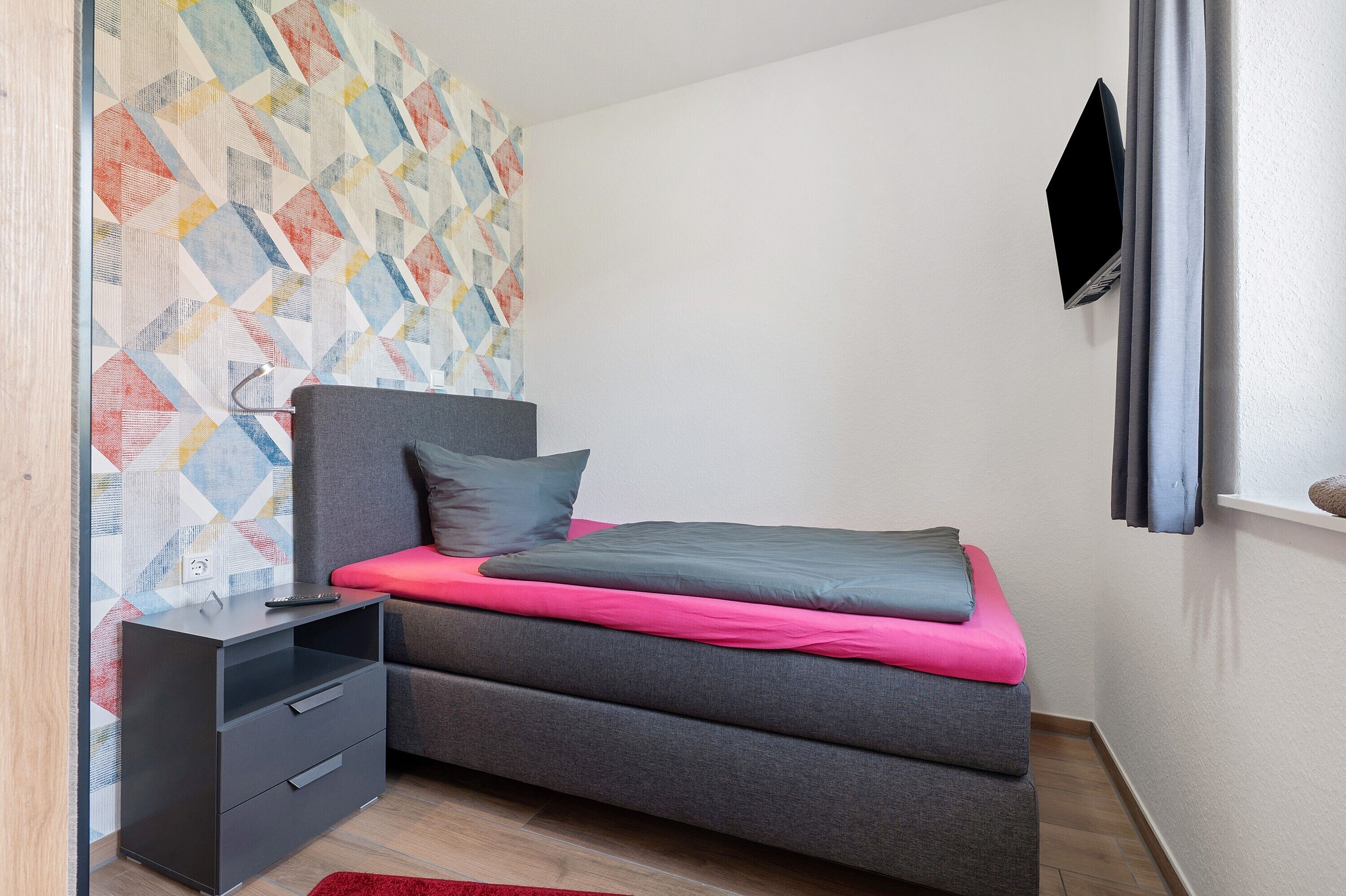 A small, modern bedroom with a bed, a nightstand, a TV mounted on the wall, and a patterned wall.