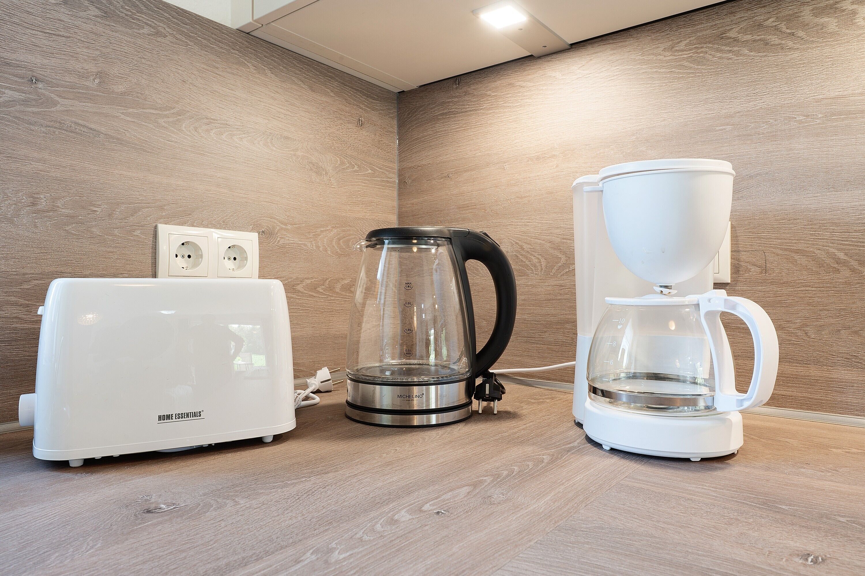 Eine Küchenzeile mit einem weißen Toaster, einer Glas-Kaffeekanne und einer weißen Kaffeemaschine auf einer Holzauslage.