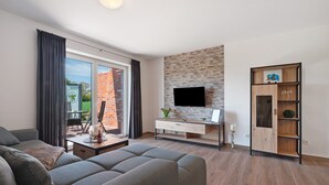 A modern living room with a grey sofa, a flat-screen TV mounted on a stone wall, a wooden cabinet with shelves, and a dining table with chairs. - Holiday Apartment 'Anna 2 Eg' with Private Terrace and Garden (Suhle)