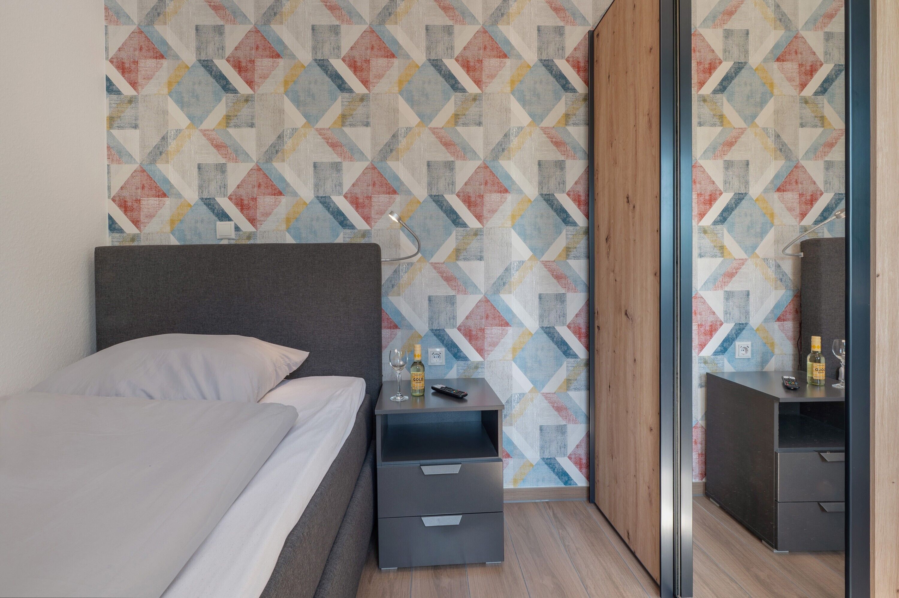 A modern hotel room with a bed, a bedside table with a remote control, a bottle of alcohol, and a mirror.