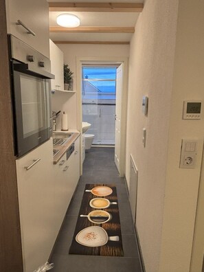 A narrow hallway with a kitchen area on one side, a toilet, and a window leading to an outdoor balcony.