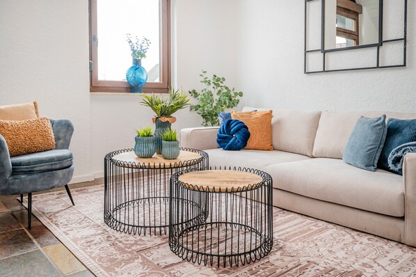 A living room with a sofa, two round coffee tables, a chair, and a window with plants.