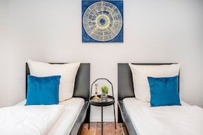 A room with two beds, each with a pillow, a small table with a plant and a vase, and a wall-mounted artwork.