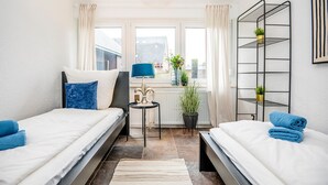 A bedroom with two beds, a small table, a radiator, a window with curtains, and a plant on a shelf.