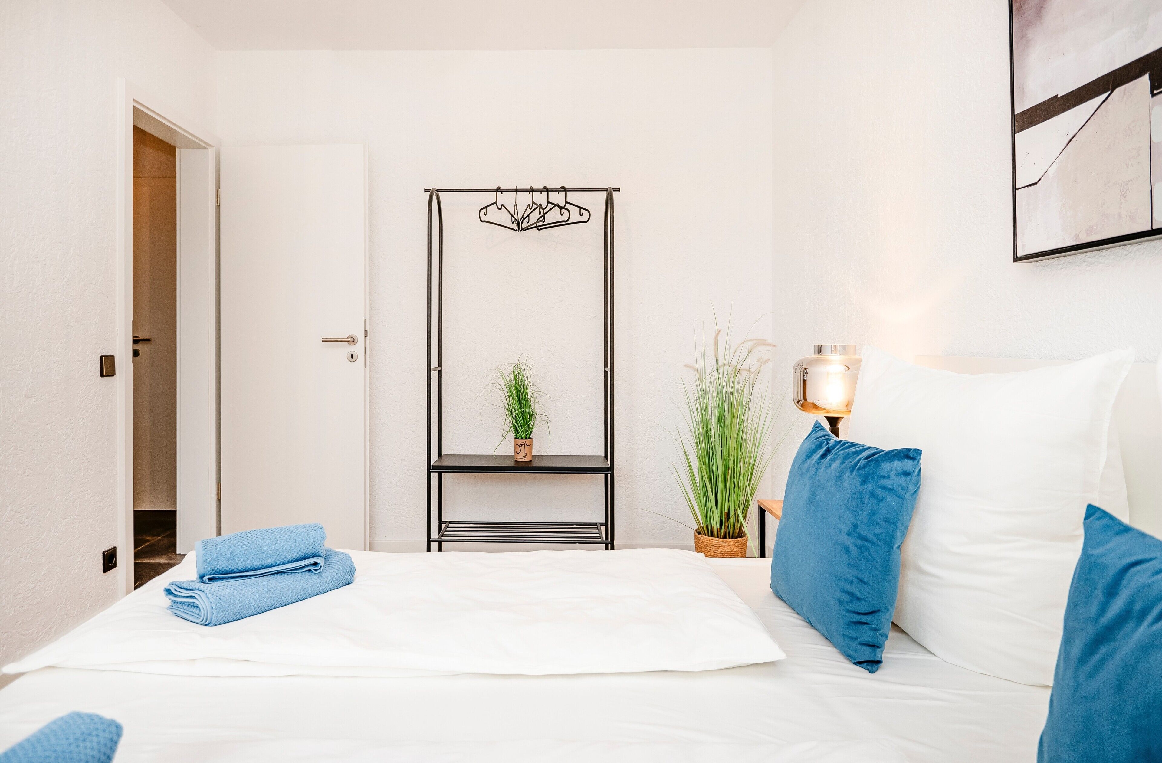 A hotel room with a bed, a bedside table with a lamp, a wall-mounted shelf with a plant, and a door leading to another room.