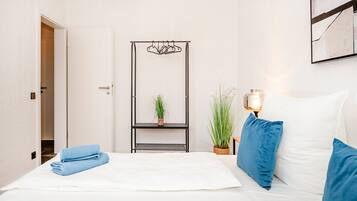 A hotel room with a bed, a bedside table with a lamp, a wall-mounted shelf with a plant, and a door leading to another room.