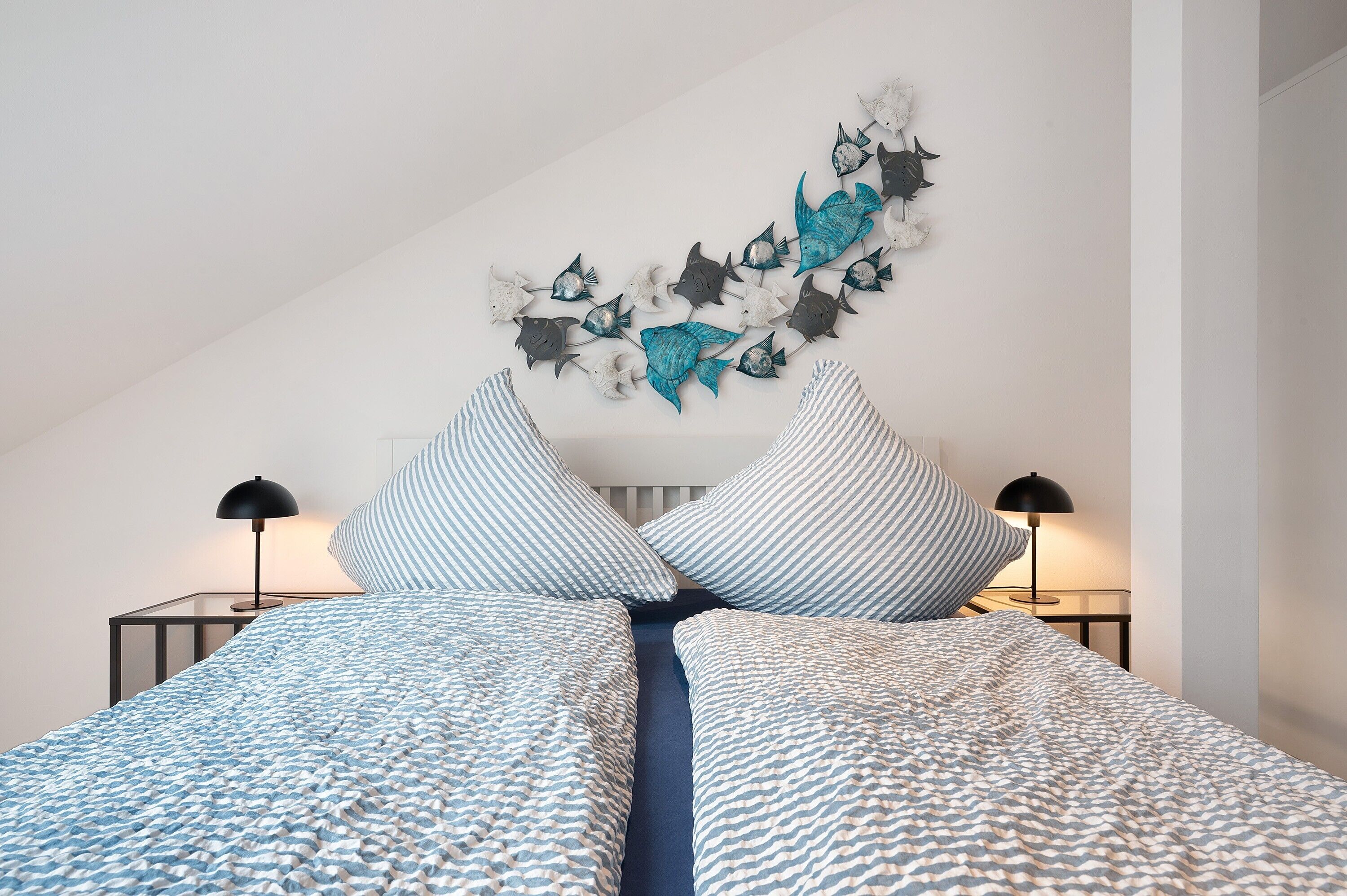 Un dormitorio con cama, almohadones a rayas, un cuadro de peces y dos lámparas de noche.