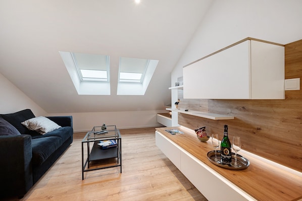 Un loft moderno con tragaluces, un sofá azul oscuro, una mesa de centro de vidrio y una encimera de madera con una botella y vasos.