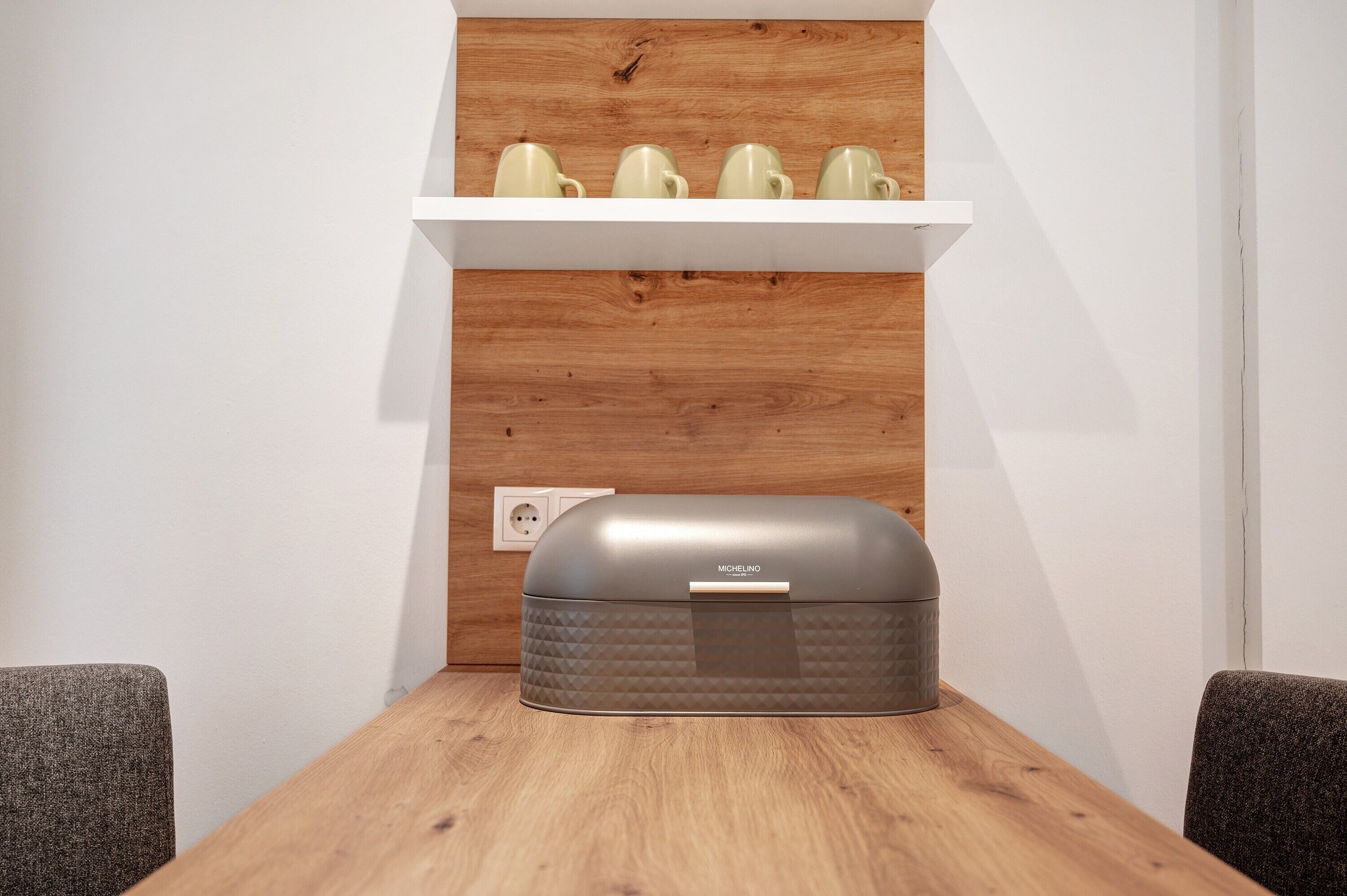Een houten tafel met een grijze opbergdoos, een witte plank met keramische mokken, en een stopcontact aan de muur.