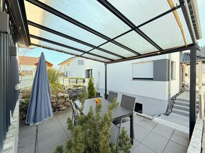A covered patio with a table and chairs, a blue umbrella, and a view of a white building with a stone wall and a wooden fence.