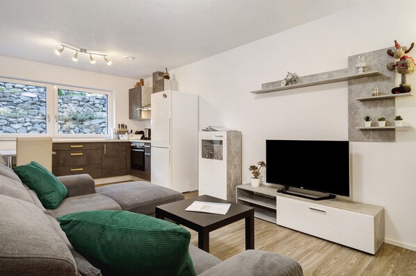 A modern living room with a grey sofa, a coffee table, a flat-screen TV, and a kitchen area with a fridge, stove, and cabinets.