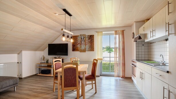 Una cocina moderna con armarios de madera, un comedor con mesa y sillas, un televisor y un balcón con cortinas.