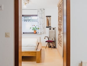 Ein Schlafzimmer mit einem hölzernen Bett, einem Nachttisch mit rotem Kissen, einem Fenster mit Vorhängen, einer Topfpflanze und einem wandmontierten Spiegel.