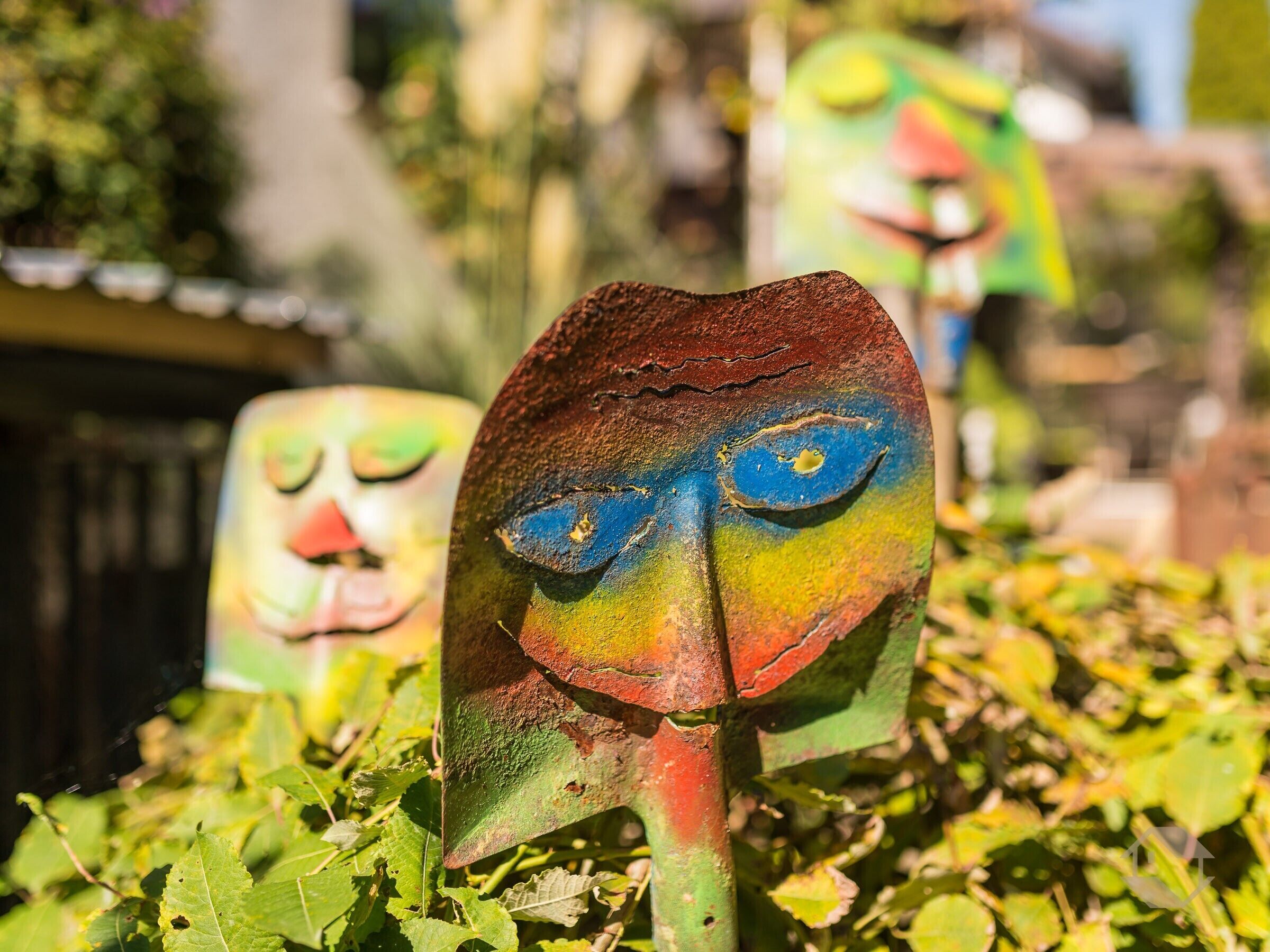 A close-up of a colorful, rustic sculpture with a face, surrounded by greenery.