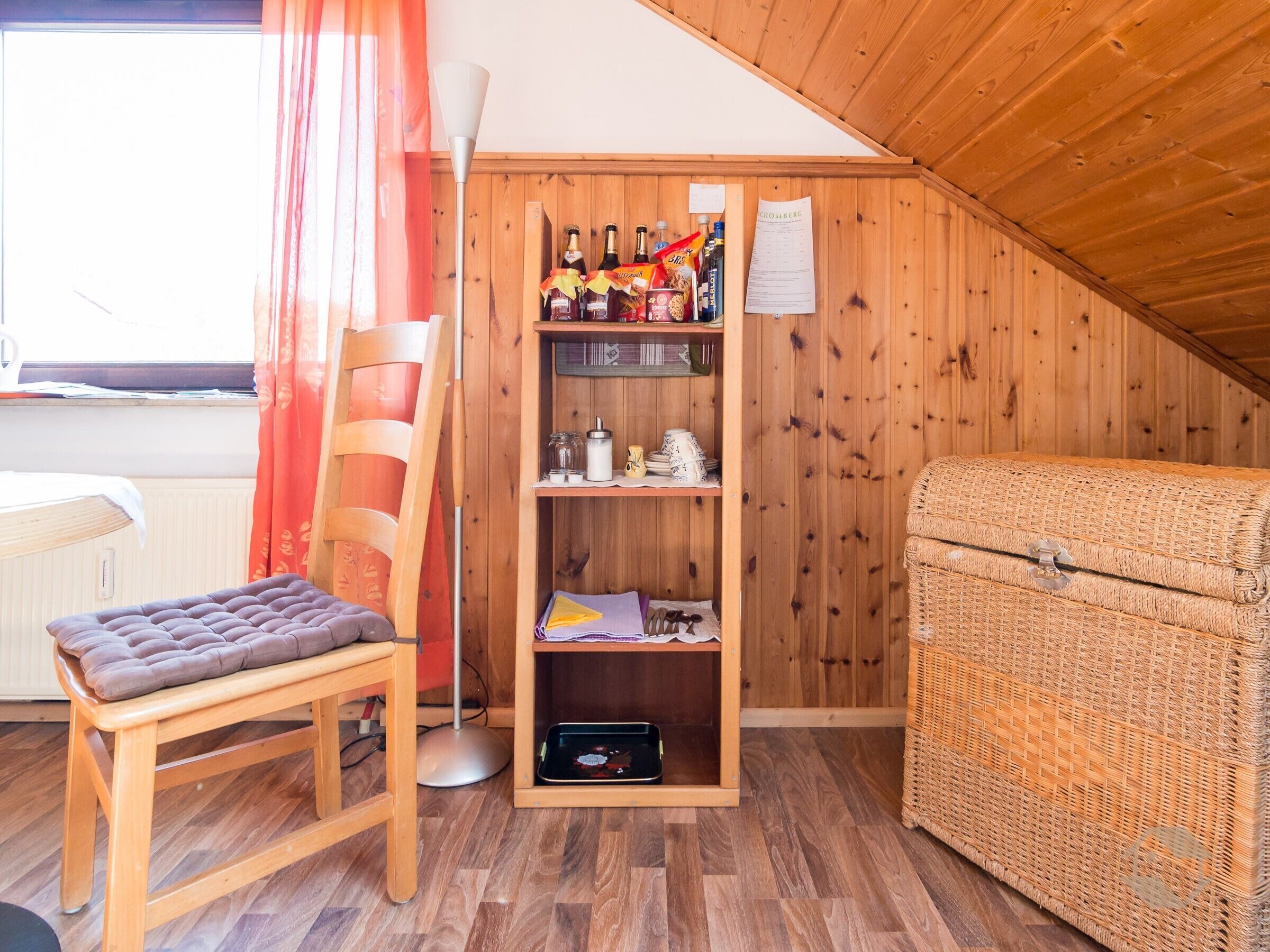 Una habitación acogedora con una estantería de madera, un sillón mullido, un cofre de mimbre y una ventana con cortinas.