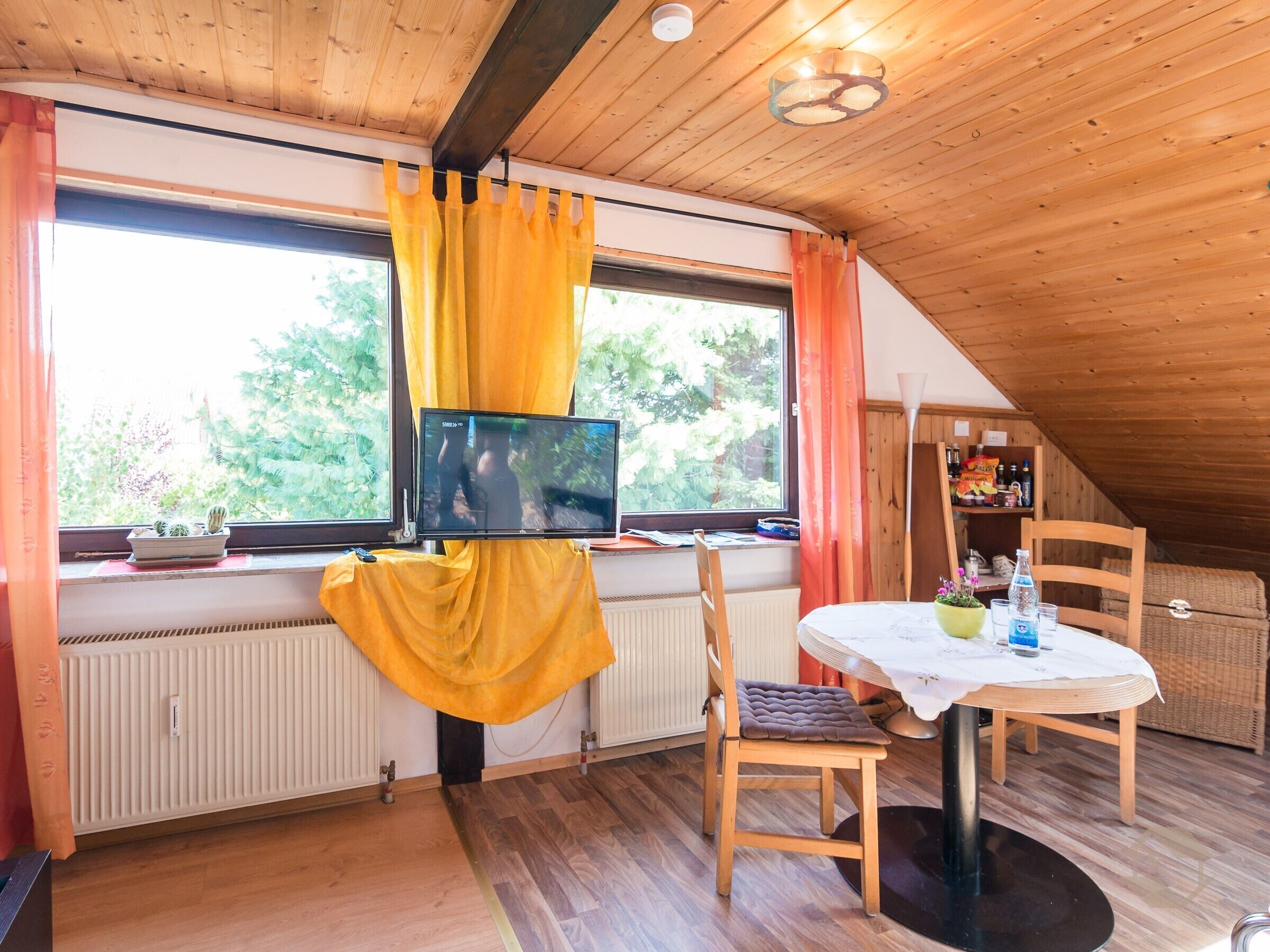 Una habitación acogedora con una televisión sobre un mueble, una mesa con sillas, un radiador y una ventana con cortinas.