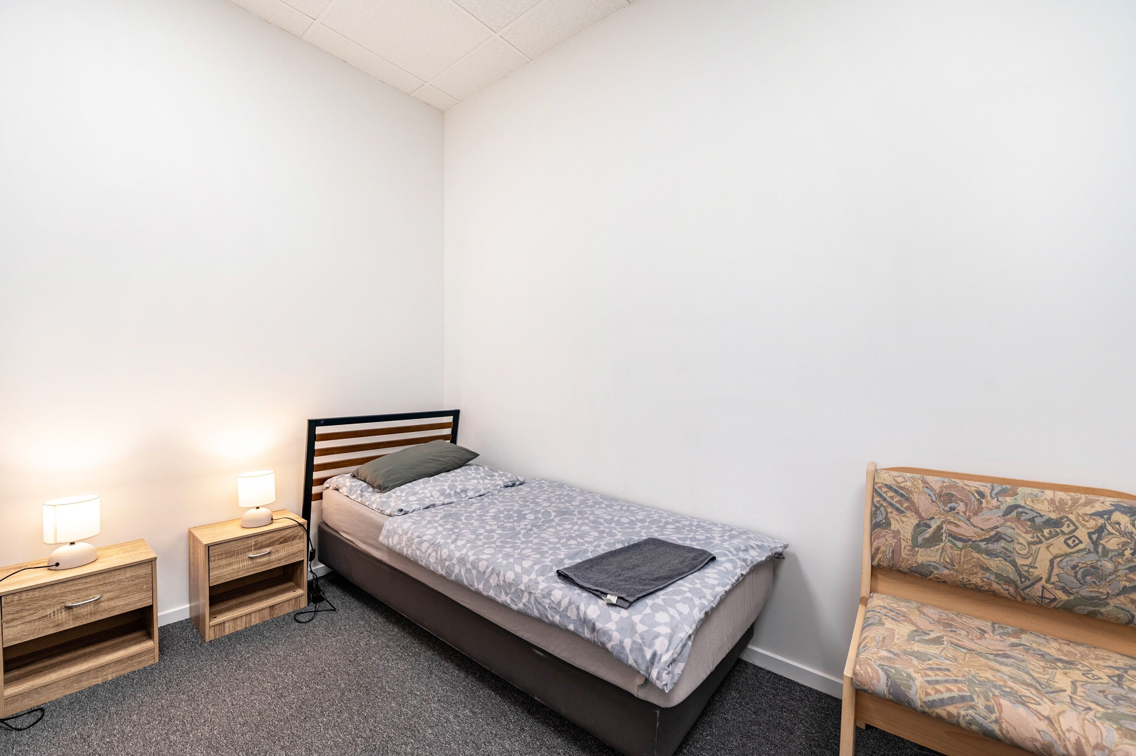 Une petite chambre à coucher, bien rangée, avec un lit simple, deux tables de chevet avec des lampes, une chaise et un canapé à motifs.