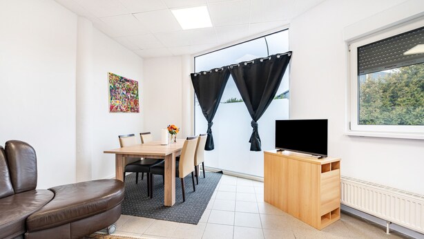 A room with a brown leather sofa, a wooden dining table with chairs, a TV on a wooden stand, and a window with curtains.