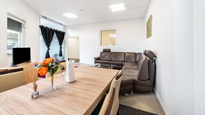 A hotel room with a wooden dining table, a brown leather sofa, a television, and a window with curtains.