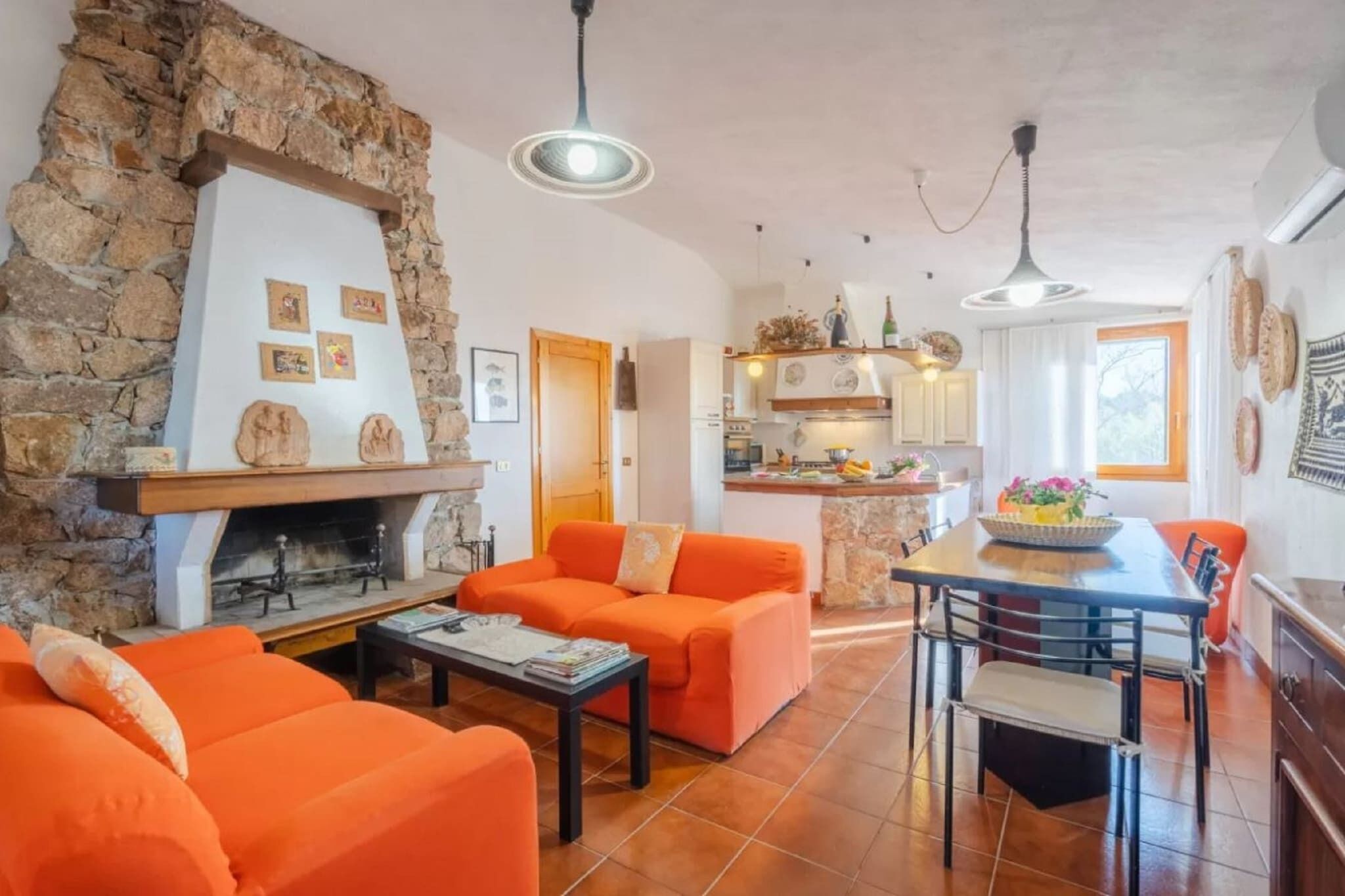 Una sala de estar con chimenea de piedra, sofás naranja, una mesa de comedor con sillas y una cocina al fondo.