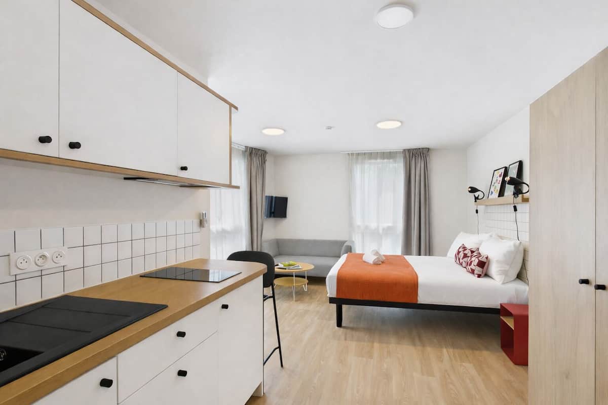 Une chambre d’hôtel compacte comprenant un coin cuisine, un lit avec une couverture orange, une télévision et une petite table.