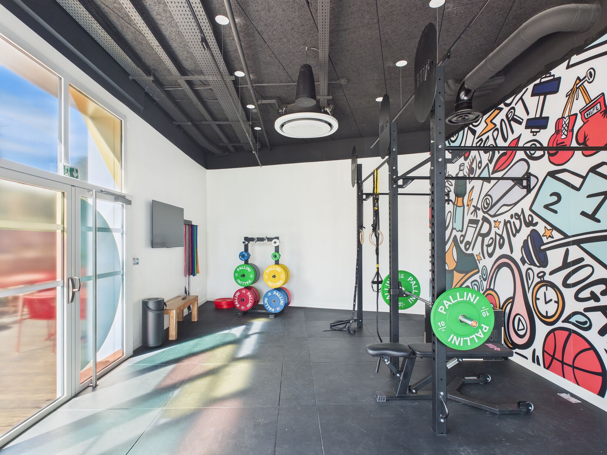 Ein Fitnessraum mit Gewichthebern, einer Wand mit Fitness-Motiven und großen Fenstern für viel Tageslicht.
