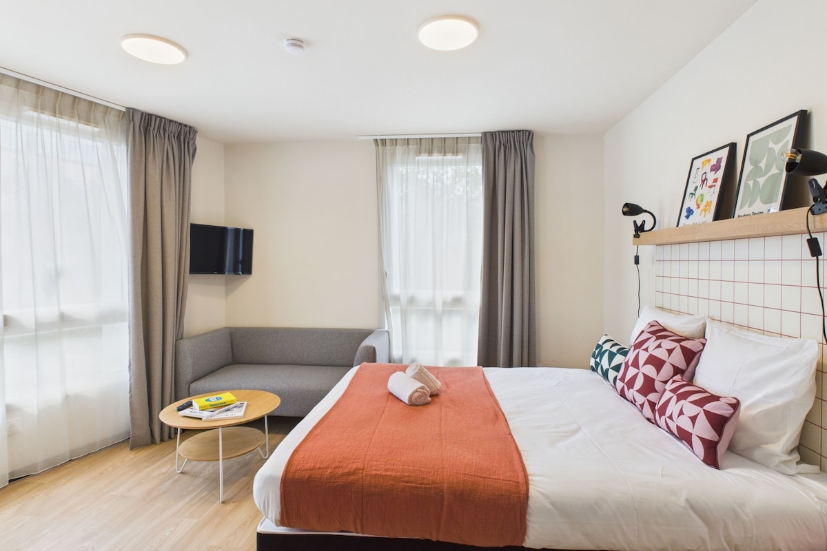 Une chambre d’hôtel avec un lit, un canapé, une petite table, une télévision et une fenêtre avec des rideaux.