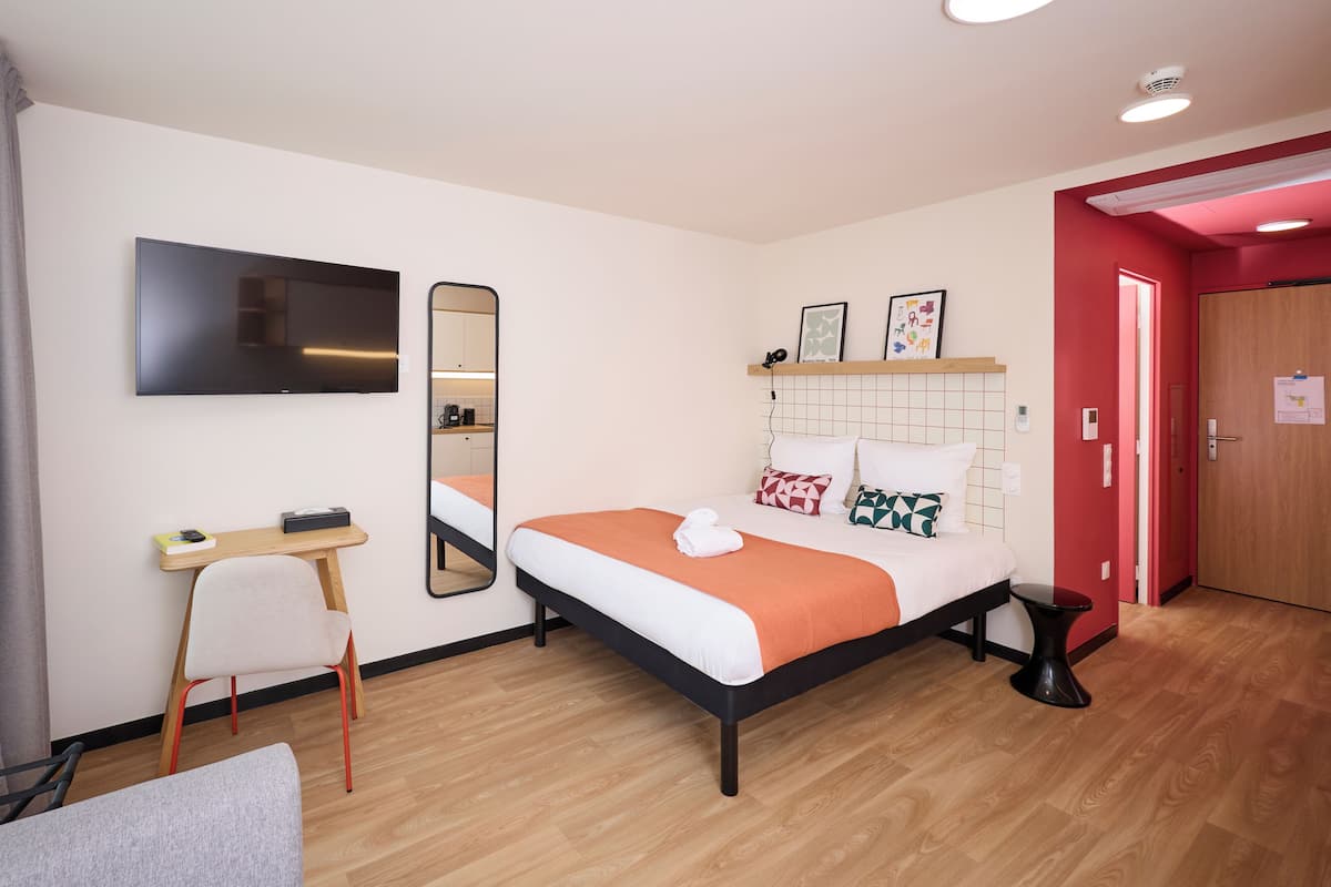 Une chambre d’hôtel moderne équipée d’un lit, d’un bureau, d’une chaise, d’une télévision, d’un miroir et d’un mur d’accent rouge.