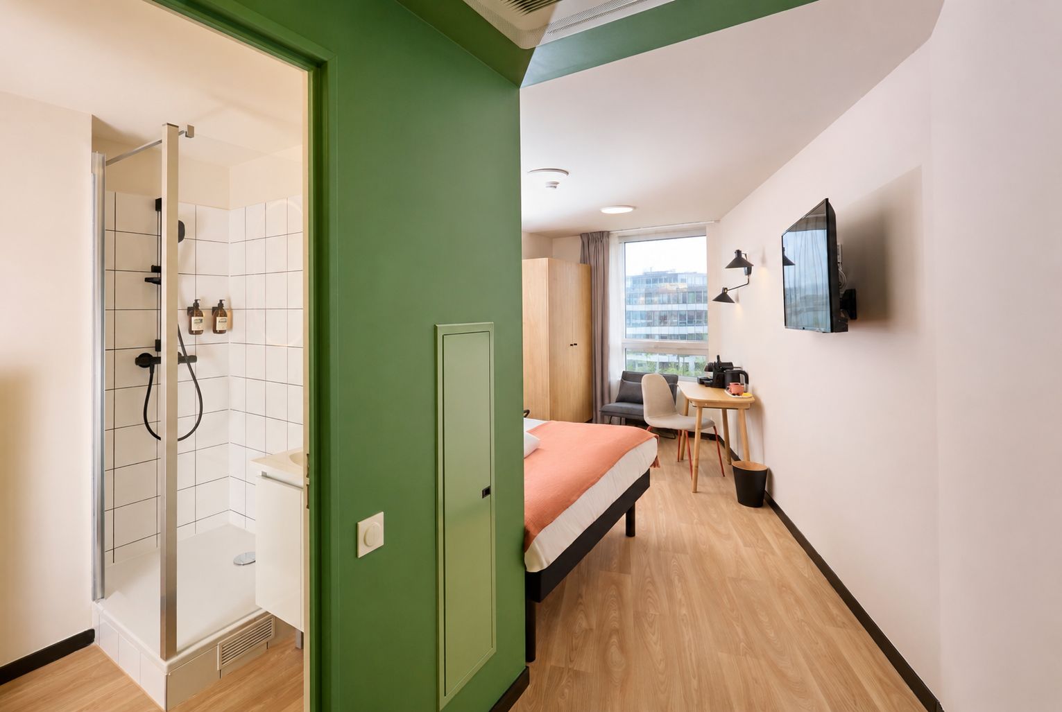 A hotel room with a shower, a bed, a desk with a chair, and a TV mounted on the wall.