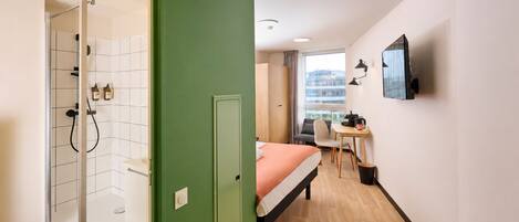 A hotel room with a shower, a bed, a desk with a chair, and a TV mounted on the wall.