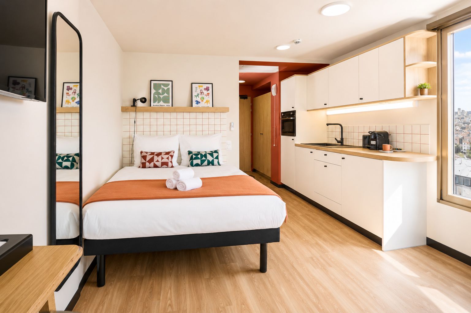 A modern hotel room with a bed, a small kitchenette, a bathroom, and a city view.