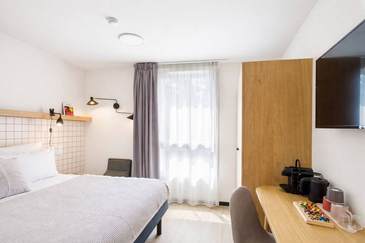 Une chambre d’hôtel moderne équipée d’un lit, d’une table de chevet, d’une télévision et d’une fenêtre avec des rideaux.