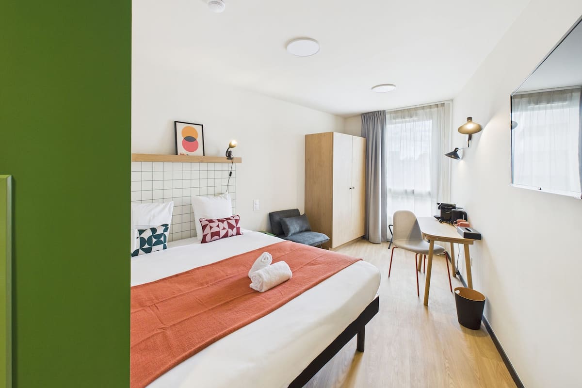 Une chambre d’hôtel avec un lit, un bureau, une chaise, une armoire et une fenêtre avec des rideaux.