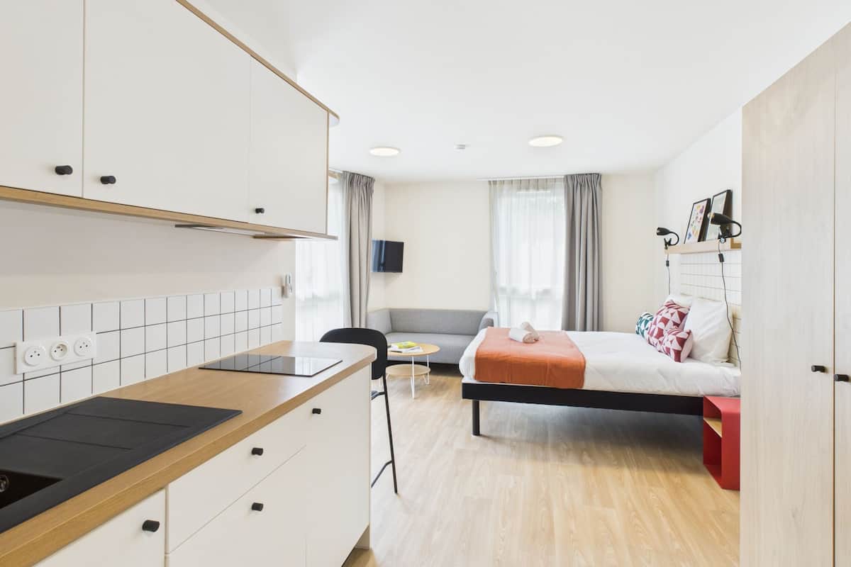 Une chambre d’hôtel moderne équipée d’une kitchenette, d’un lit agrémenté d’une touche de couleur rouge, d’une télévision et d’une fenêtre avec des rideaux.