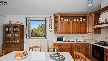 Ett kök med träskåp, ett matbord med frukt på och ett fönster med utsikt över träd.