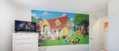 A child's bedroom with a large wall mural, a TV, and two beds with Mickey Mouse bedding.