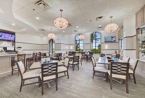 A hotel dining area with multiple tables and chairs, ceiling lights, and a television screen.