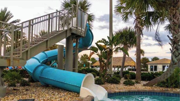 Un tobogán de agua que desemboca en una piscina rodeada de palmeras y un edificio con techo de paja.