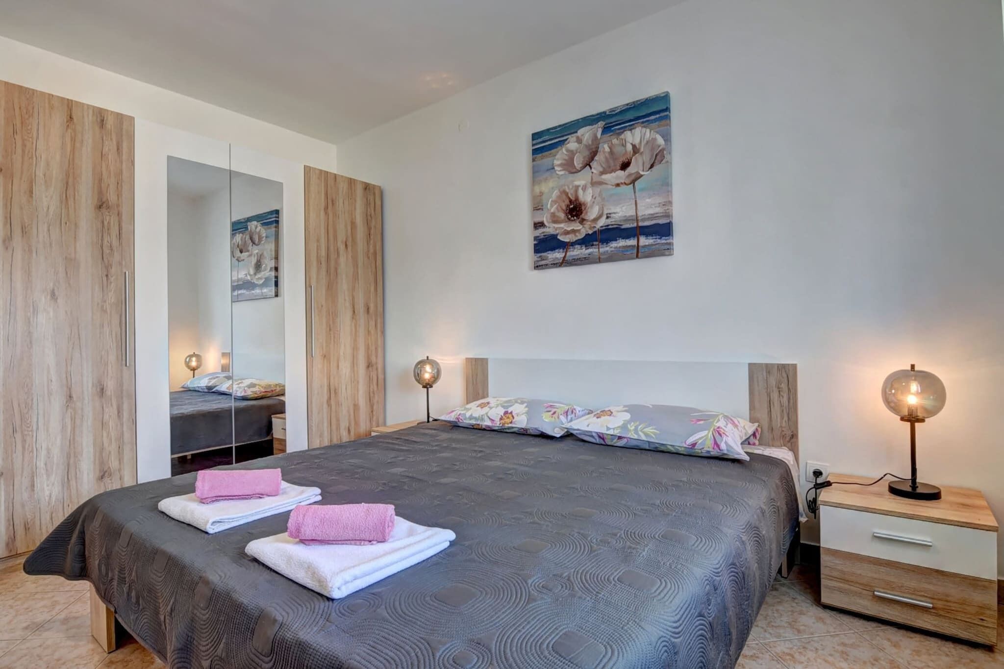 A bedroom with a large bed, a bedside table with a lamp, a mirror, and a painting on the wall.