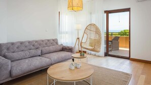 A modern living room with a grey sofa, a round wooden coffee table, a wicker chair, and a large window with a view of the outdoors.