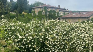 Un bâtiment en pierre, surmonté d’un toit de tuiles, entouré d’une végétation luxuriante et d’arbustes à fleurs blanches.