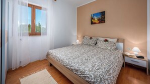 Un dormitorio con cama, mesita de noche, ventana con cortinas y un cuadro en la pared.