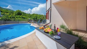 Una piscina con sombrilla, una mesa con frutas y bebidas, y vistas a colinas verdes.