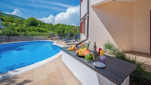 A swimming pool with a sun umbrella, a table with fruits and drinks, and a view of green hills.